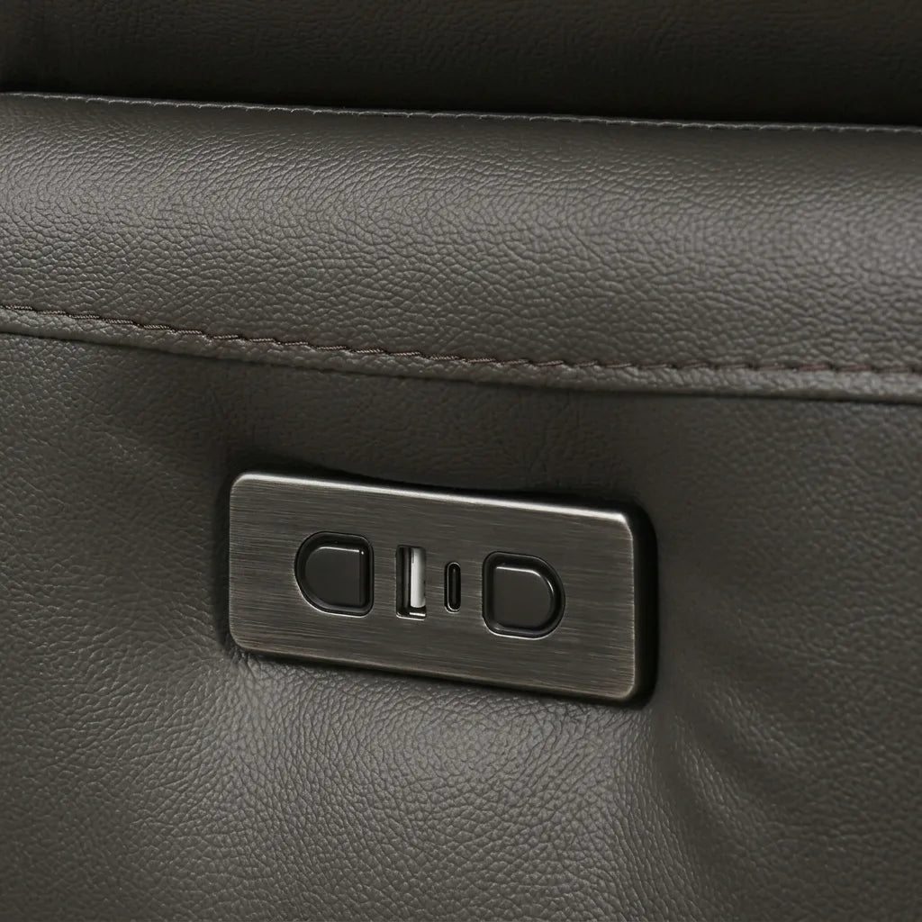Close-up of the grey-brushed rectangular power control panel for a grey leather recliner. The panel features two control buttons and an integrated USB-A charging port. The surrounding surface is grey pebbled leather.