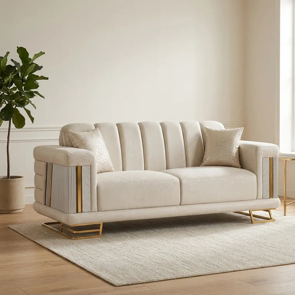 A cream-colored upholstered loveseat with vertical channel tufting on the back, fluted side panels, and gold metal legs, styled in a modern living room setting with a rug, side table, and potted plant.