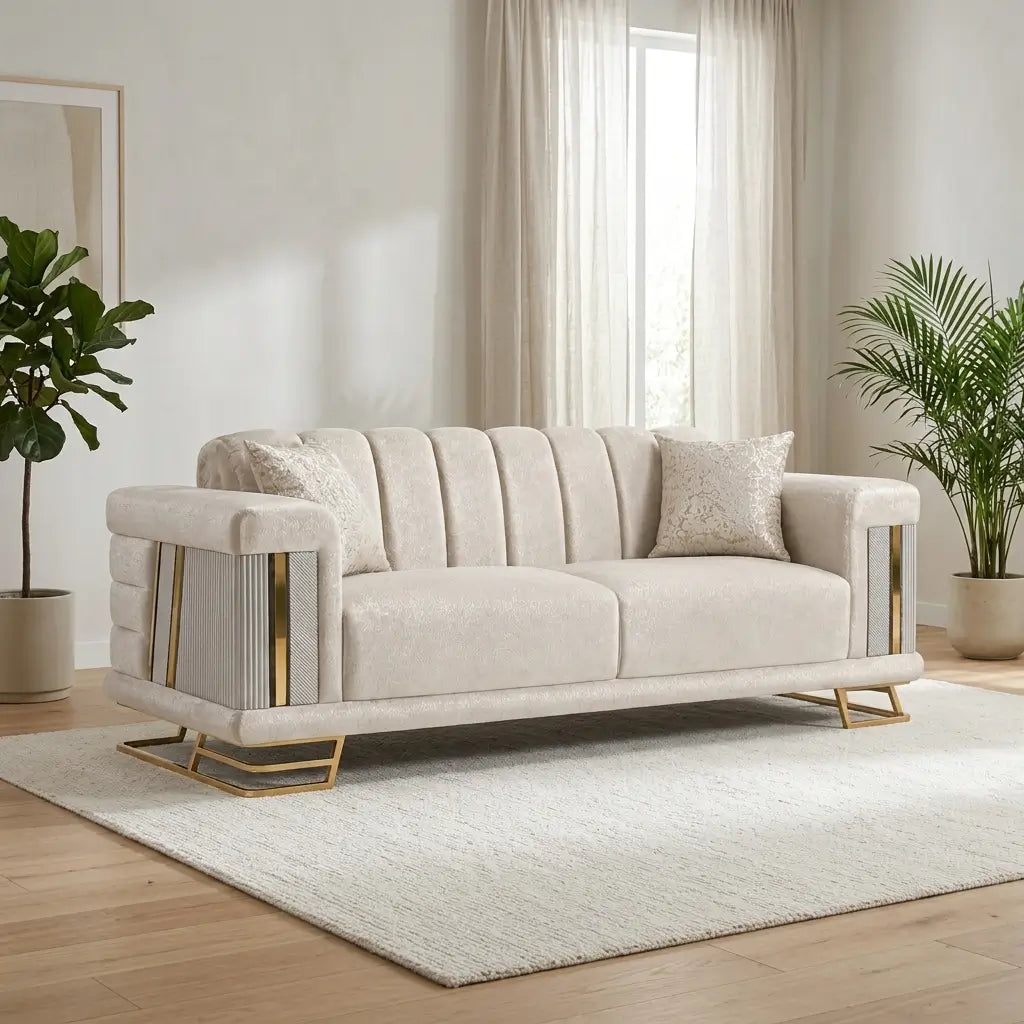 A cream-colored upholstered loveseat sofa with vertical channel tufting, gold metal side accents, and gold geometric legs, styled in a bright living room on a white rug.