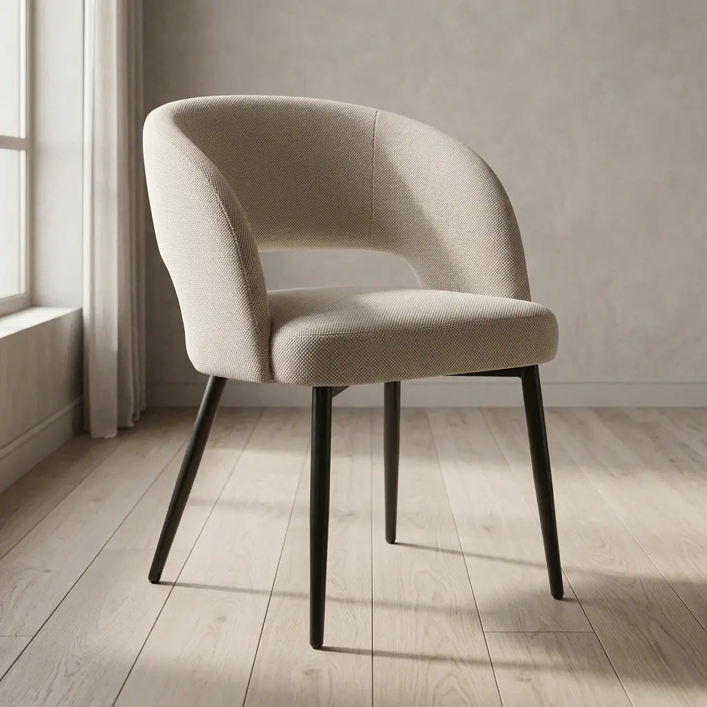 A modern barrel-style dining chair upholstered in textured beige fabric with black tapered legs, shown in a bright room with light wood flooring and a textured neutral wall.