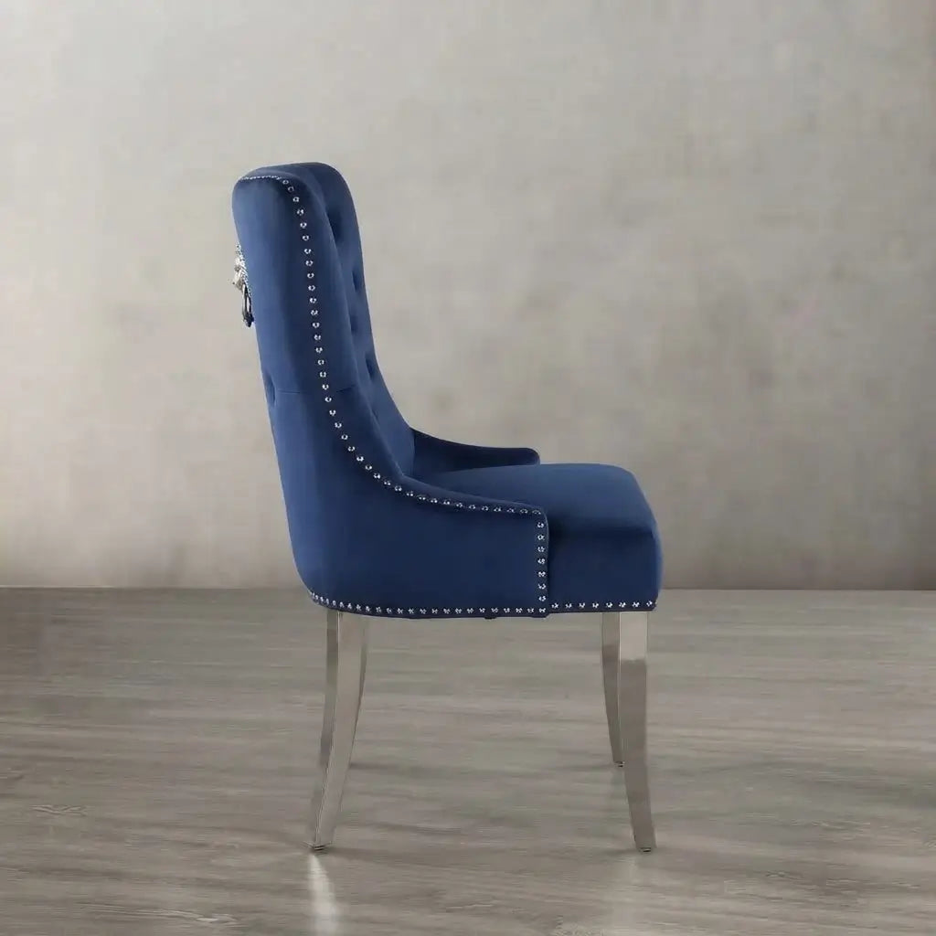 A royal blue velvet dining chair with silver nailhead trim and a silver metal leg, shown from a side profile against a neutral gray textured wall and floor.