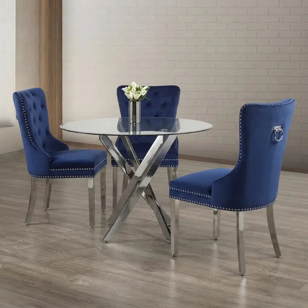 A 4-piece dining set featuring a round glass-top table with a chrome X-base and three blue tufted velvet chairs with silver nailhead trim and ring pulls, set in a dining room with a light brick wall and wood floor.