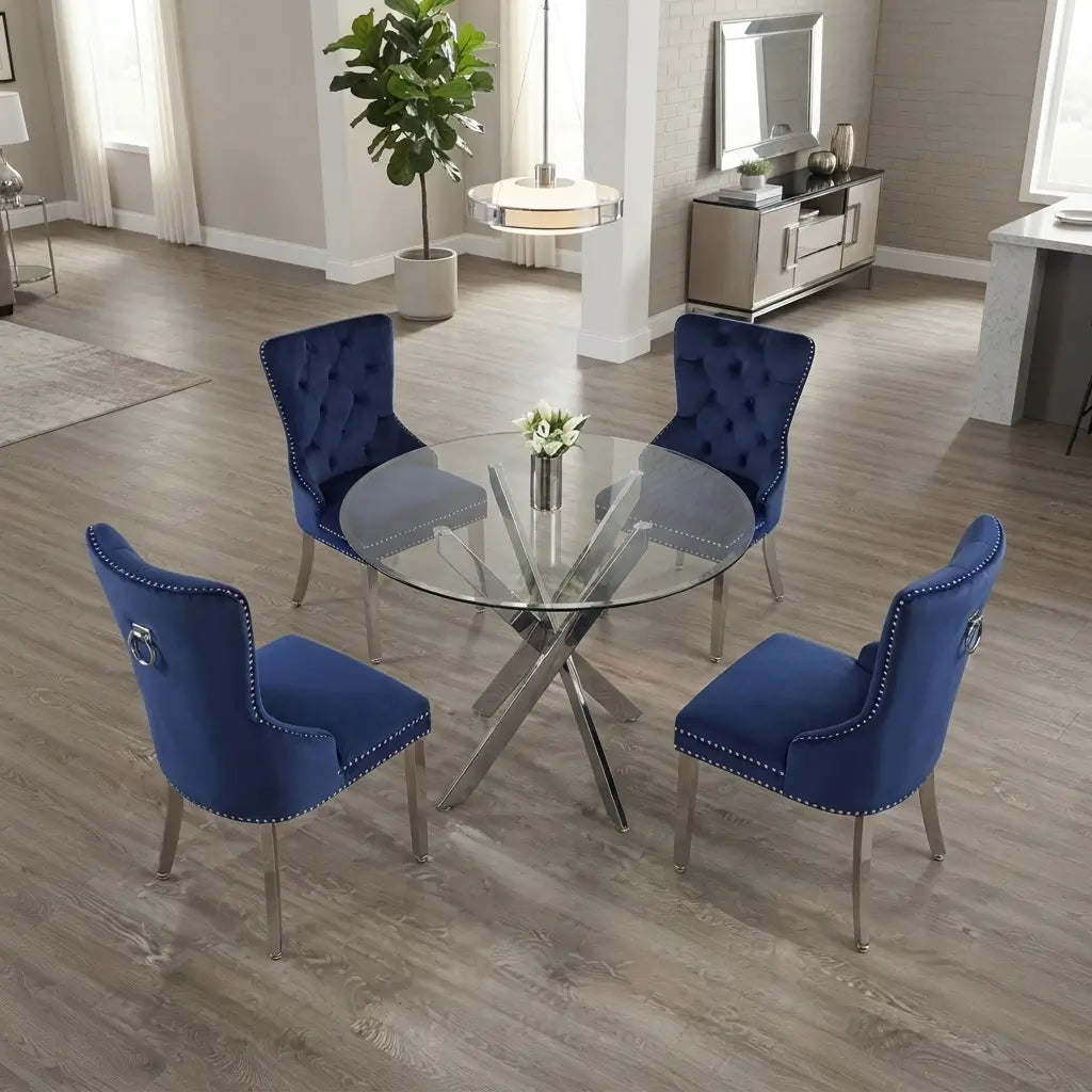 A 4-piece dining set featuring a round glass-top table with a chrome X-base and four tufted blue velvet chairs with silver nailhead trim and chrome legs, shown in a modern dining room setting.