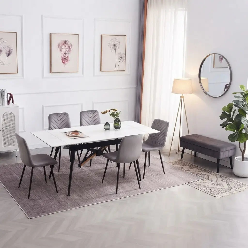 A modern dining room setup featuring a rectangular white marble-top dining table with a black metal frame and six gray upholstered chairs, styled in a bright, contemporary room with a floor lamp, bench, and potted plant.