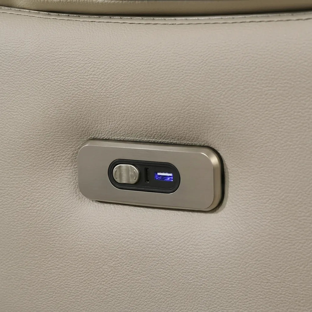 A close-up photograph displays a metal control panel with a rounded rectangular form, featuring a satin champagne-gold finish. The panel is set flush into a beige, finely textured leather material that is soft to the touch. A silver rocker switch with a fine mesh pattern is located on the left side, and a standard rectangular USB type-A port on the right. In the center of the USB port, a small internal indicator light emits a blue glow, highlighting its functionality and ready state. Detailed stitching runs