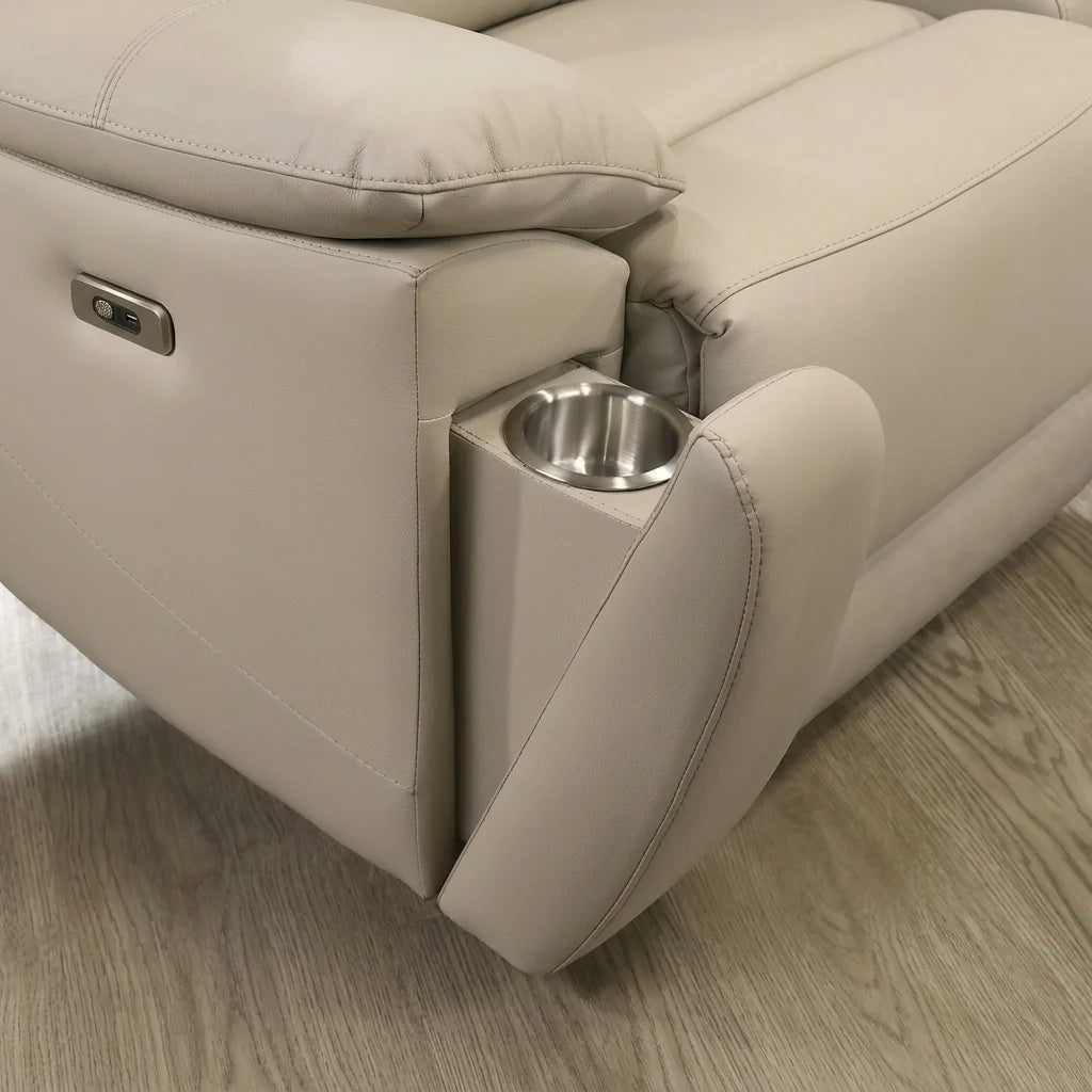 A high-definition, 1:1 ratio photorealistic close-up of the side controls on a light beige leather power recliner. A rectangular metal control panel is flush-mounted into the leather, featuring a specific round textured button and a smaller toggle-style switch. The texture of the light beige leather is clearly visible, set against the blurred light wood floor.