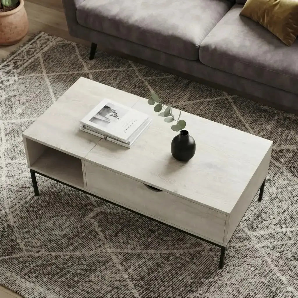 A modern rectangular coffee table with a light beige wood-grain finish and black metal legs, styled in a living room setting on a patterned rug next to a gray sofa.