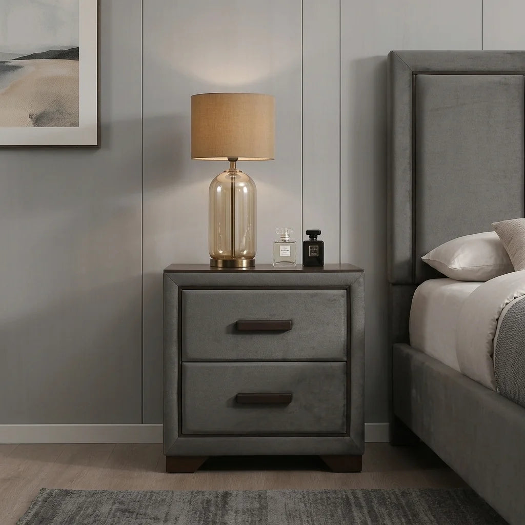 Grey upholstered 2-drawer nightstand with dark wood handles and a warm table lamp next to a padded bed headboard.
