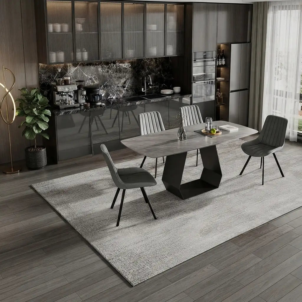 A modern dining room scene featuring a rectangular gray stone-look table with a sculptural black pedestal base, surrounded by four gray upholstered chairs on a large light gray rug. The background shows a contemporary kitchen with dark cabinets, a marble backsplash, and a refrigerator.