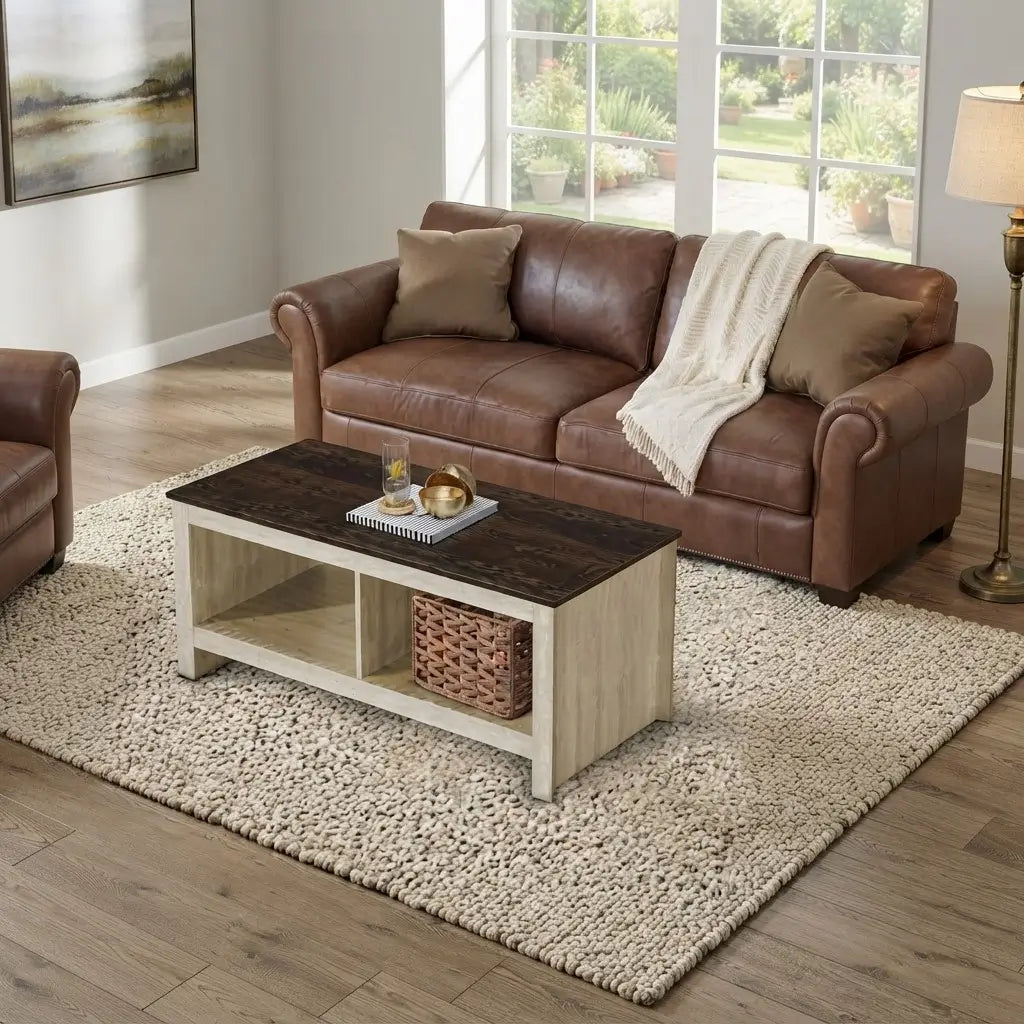 A brown leather loveseat sofa in a bright living room, styled with two tan pillows and a cream throw blanket, positioned on a large textured beige rug next to a wooden coffee table.