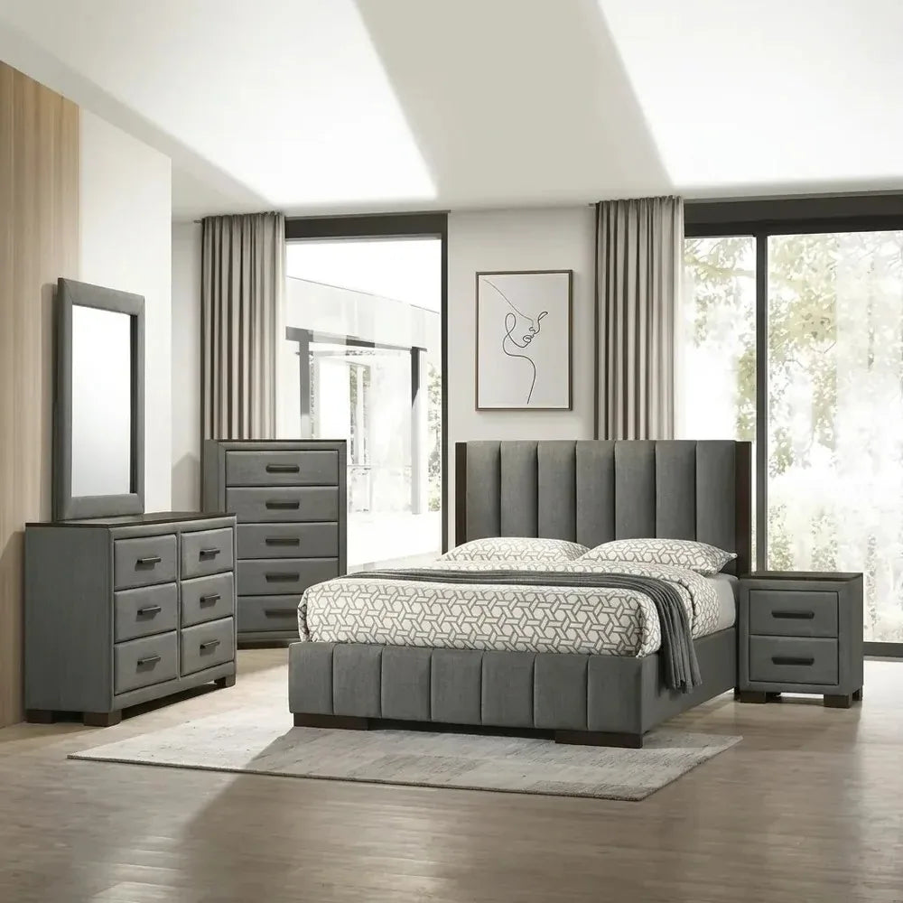A detailed view of the modern Dylan Bedroom Set, featuring a grey, vertically channeled fabric bed frame, a matching upholstered nightstand, and grey storage chests. The bed is dressed with a geometrically patterned duvet and pillows, resting on a rug in a bright room. Background elements include large windows, curtains, wood-paneled walls, and minimalist framed art.