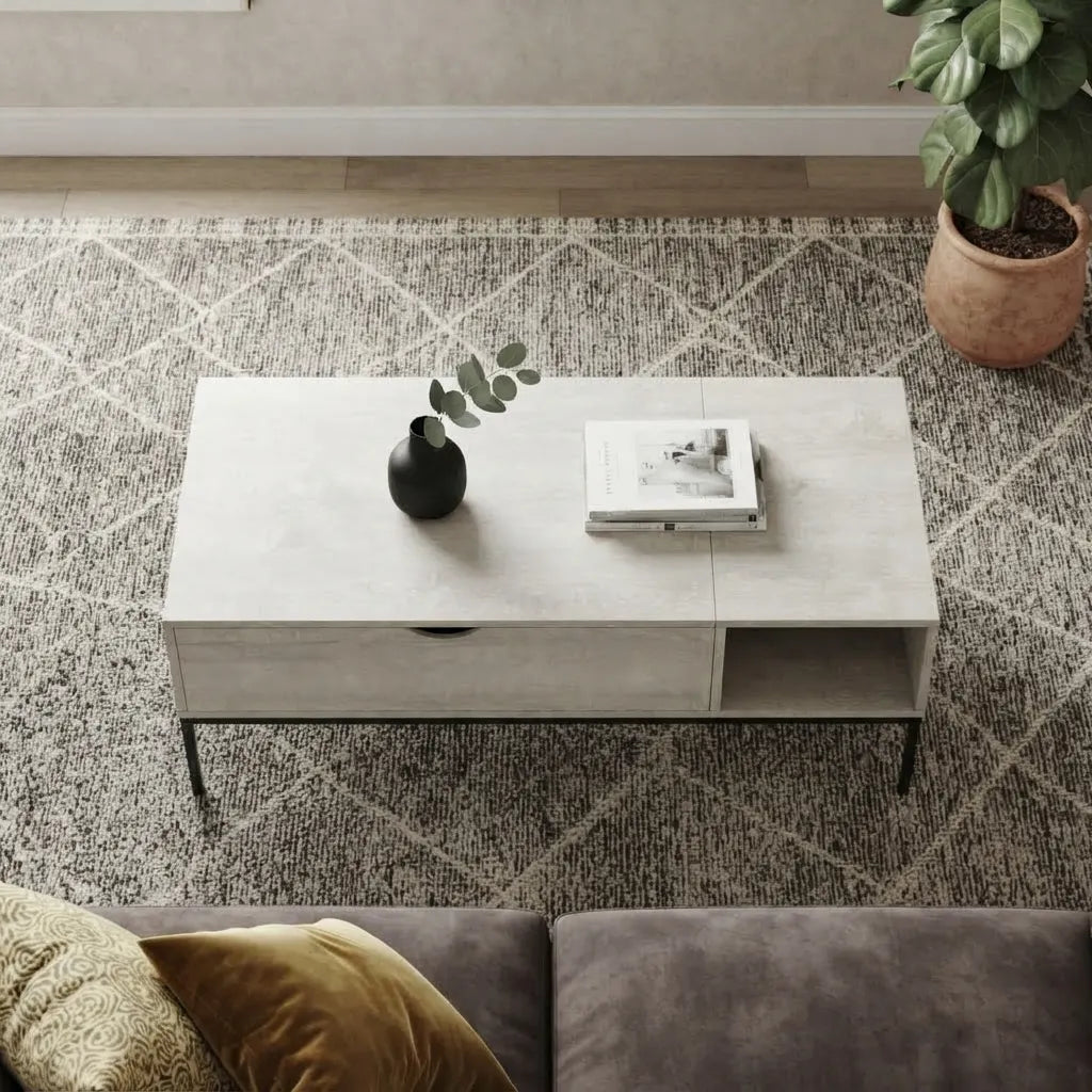 High-angle front view of a modern light oak coffee table displaying its front storage drawer and open side shelf, placed on a grey patterned rug in front of a dark velvet couch with accent pillows.