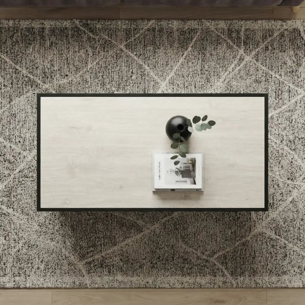 Flat-lay overhead view showing the rectangular tabletop of a light oak wood coffee table styled with a black vase containing eucalyptus leaves and a coffee table book, set on a dark textured diamond-pattern rug.